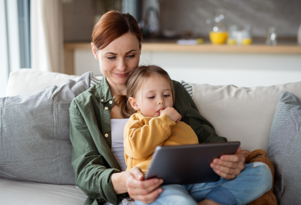 A happy mother with small daughter watching kids programme on tablet indoors at home, single parenting concept.