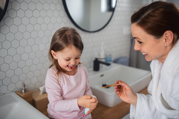 Mother with happy small daughter brushing teeth indoors in bathroom in the evening or morning.