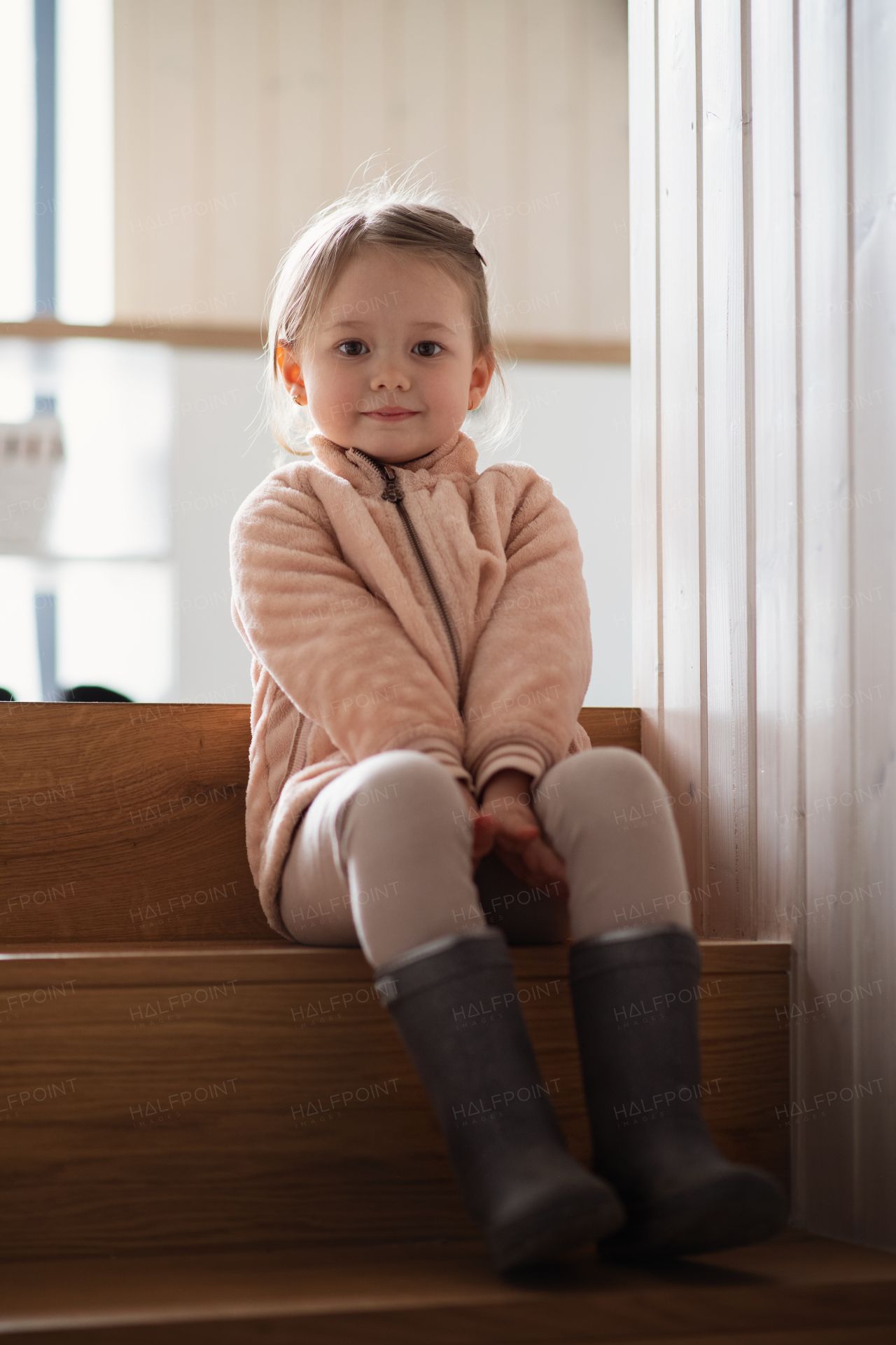 A happy small girl sitting on stairs indoors at home, ready to go out, looking at camera.