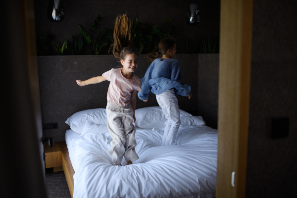 Two little sister jumping on a bed indoors in hotel.