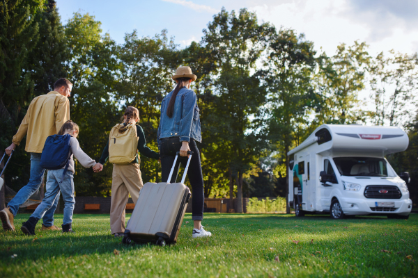 A rear view of young family with suitcases going to caravan outdoors at park.