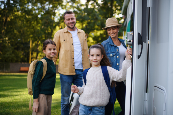 A happy young family with luggage looking at camera with camper at background outdoors in garden.