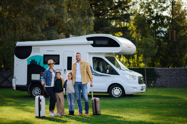 A happy young family with luggage looking at camera with camper at background outdoors in garden.