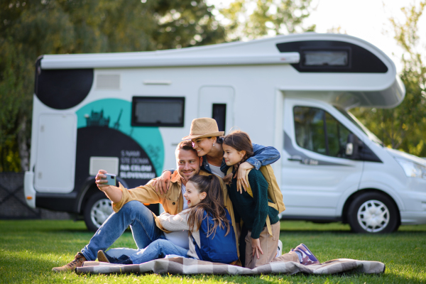 A happy young family with two children ltaking selfie with caravan at background outdoors.