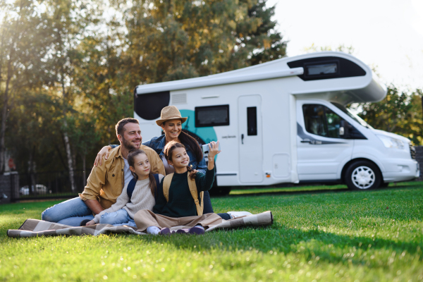 A happy young family with two children ltaking selfie with caravan at background outdoors.