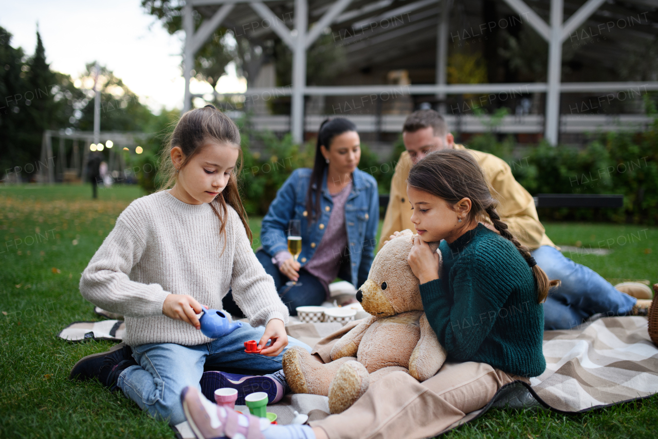 Happy little sisters sitting on blanket and playing with teddy bear on picnic with parents outdoor in park