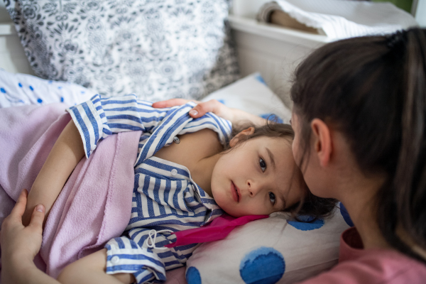 Worried mother looking after sick small daughter in bed at home, coronavirus concept.