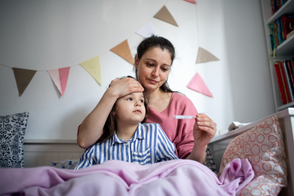 Portrait of mother looking after sick small daughter in bed at home, measuring temperature.