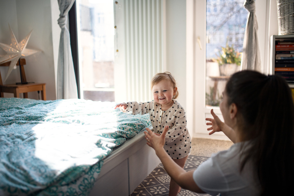 A small toddler girl with mother learning to walk in bedroom at home, first steps concept.