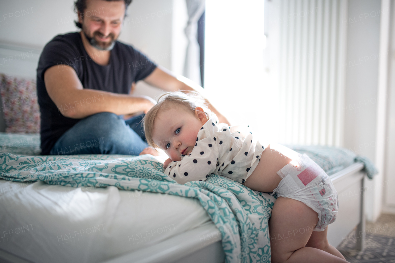 A small toddler girl with father indoors in bedroom at home, looking at camera.