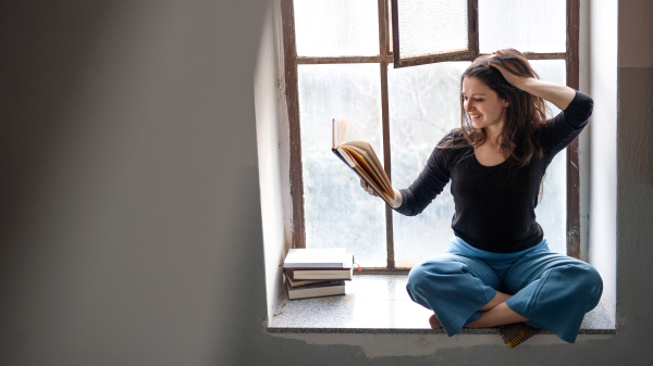 Portrait of happy woman sitting on old and dirty window sill, reading a book.