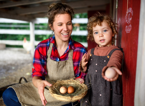 Portrait of mother with happy small daughter standing on farm, holding basket with eggs.