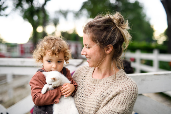 Happy small girl with mother standing on farm, playing with cat.