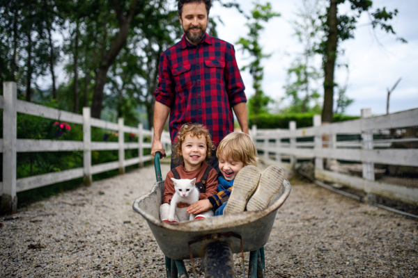 Unrecognizable father pushing cheerful small children in wheelbarrow on farm.