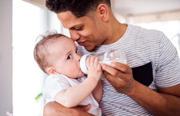 A portrait of cheerful father bottle feeding a small toddler son indoors at home.