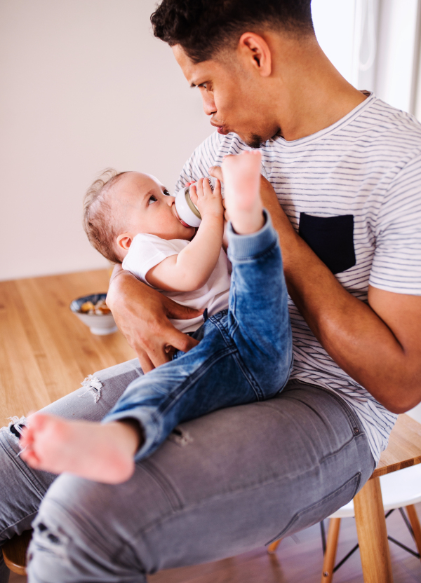 A portrait of cheerful father bottle feeding a small toddler son indoors at home.