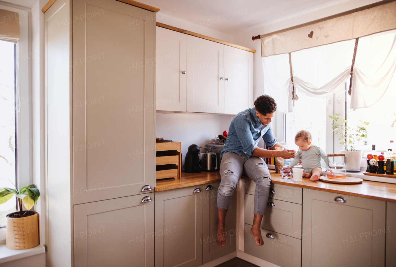 A father and a small toddler son eating fruit and yoghurt in kitchen indoors at home.