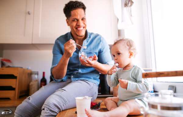 A father and a small toddler son eating fruit and yoghurt in kitchen indoors at home.