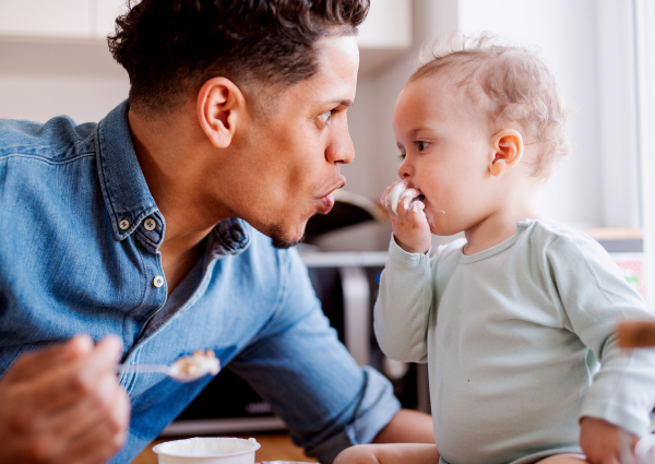 A father and a small toddler son eating fruit and yoghurt in kitchen indoors at home.