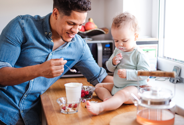 A father and a small toddler son eating fruit and yoghurt in kitchen indoors at home.