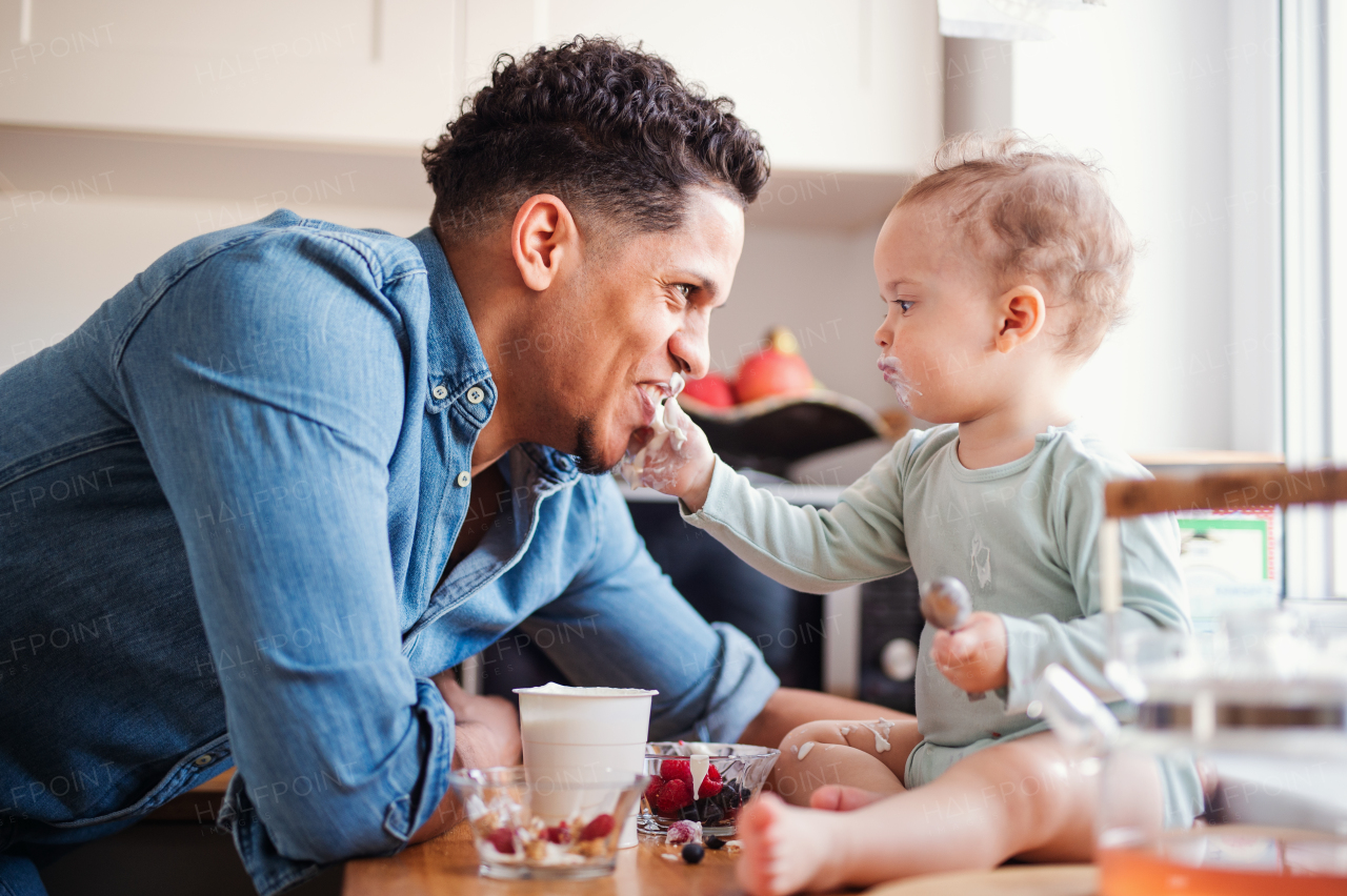 A father and a small toddler son eating fruit and yoghurt in kitchen indoors at home.