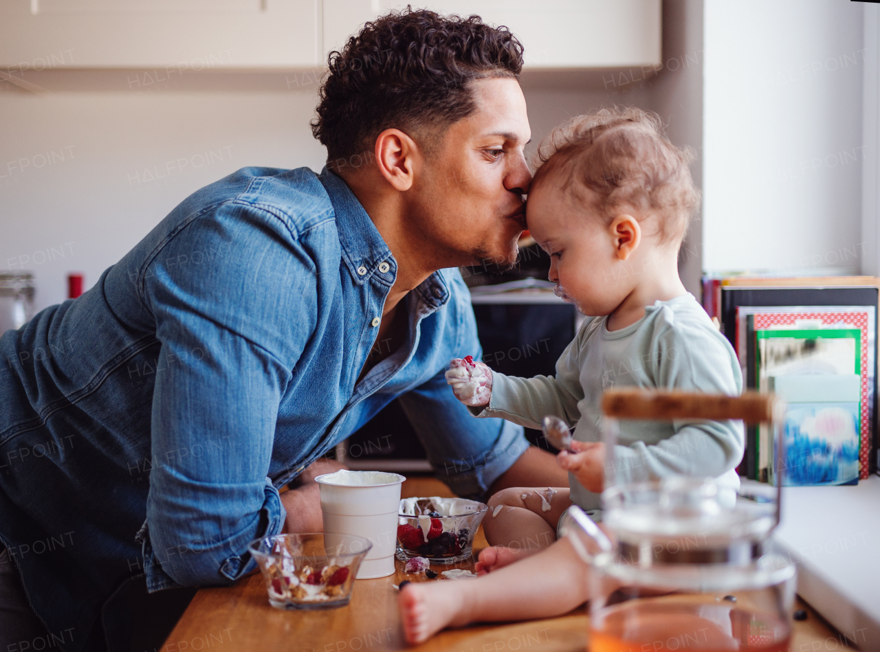 A father and a small toddler son eating fruit and yoghurt in kitchen indoors at home.