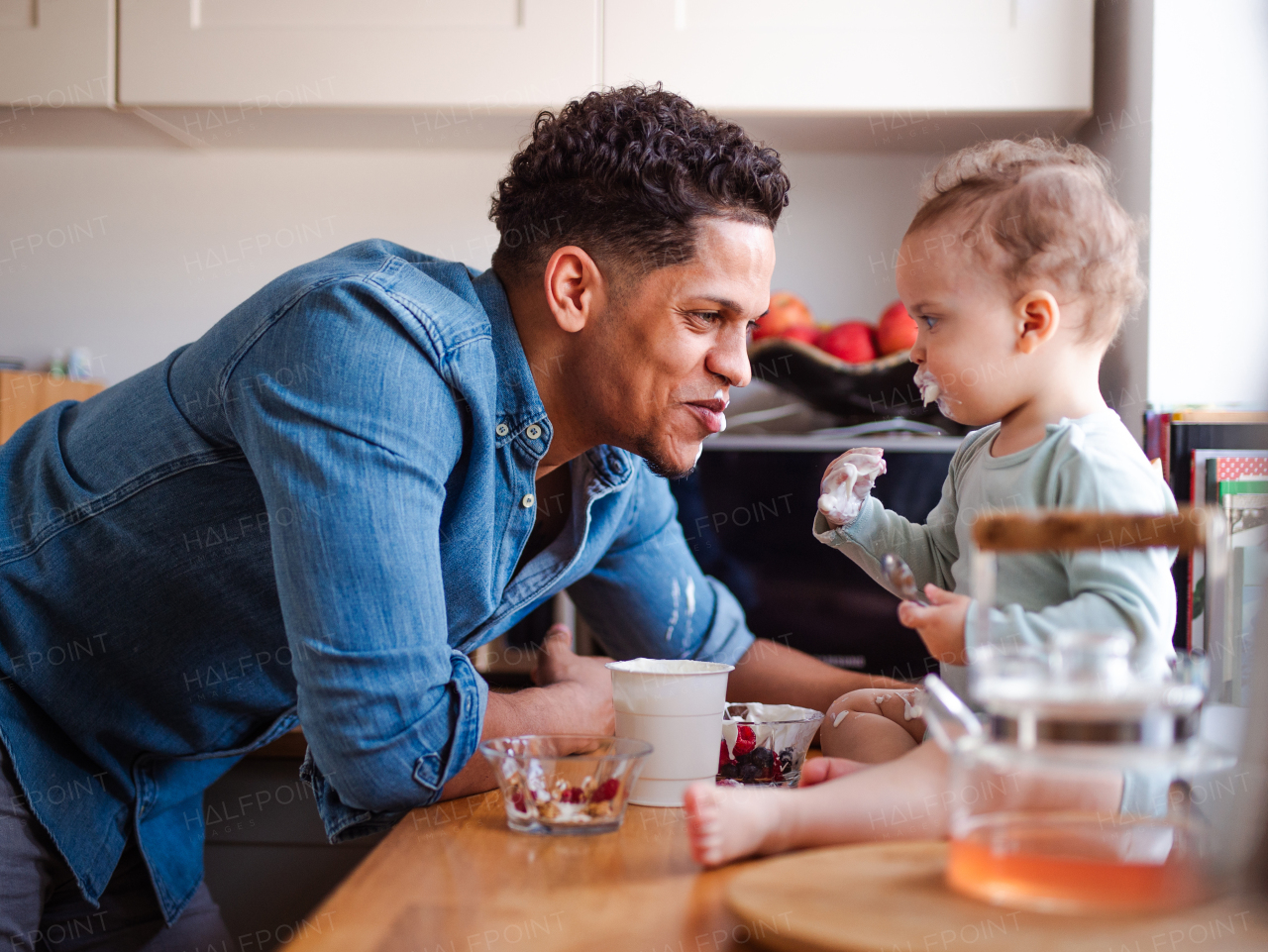 A father and a small toddler son eating fruit and yoghurt in kitchen indoors at home.