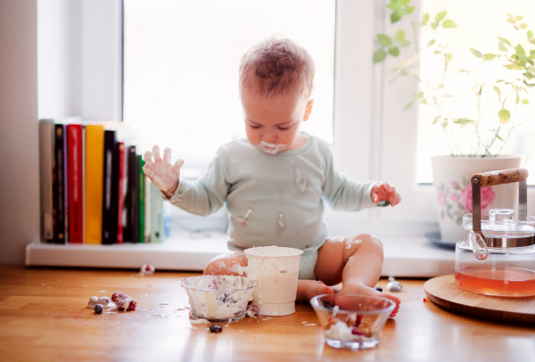 A small messy toddler boy sitting on kitchen counter at home, eating fruit and yoghurt.