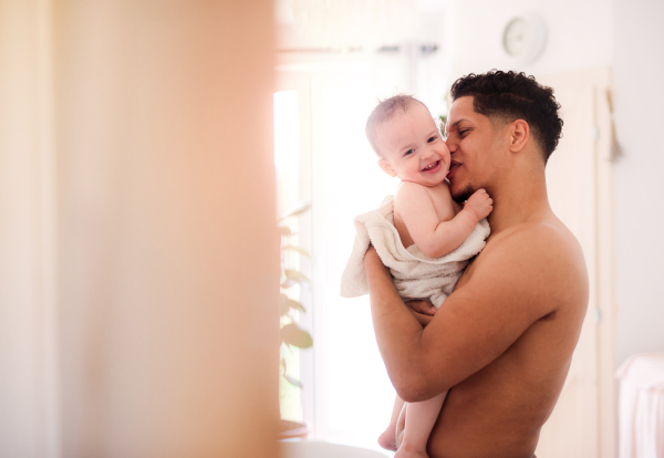 Topless father and small toddler son wrapped in towel in a bathroom indoors at home.