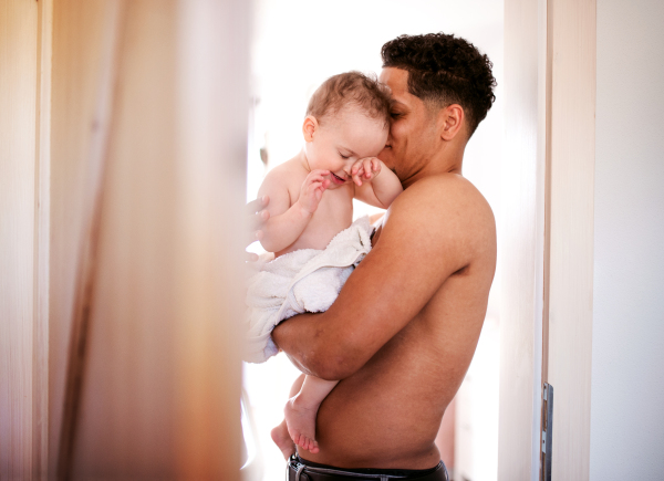 Topless father and small toddler son wrapped in towel in a bathroom indoors at home.