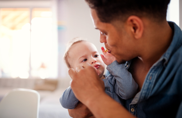 A portrait of cheerful father and small toddler son indoors at home, eating snacks.