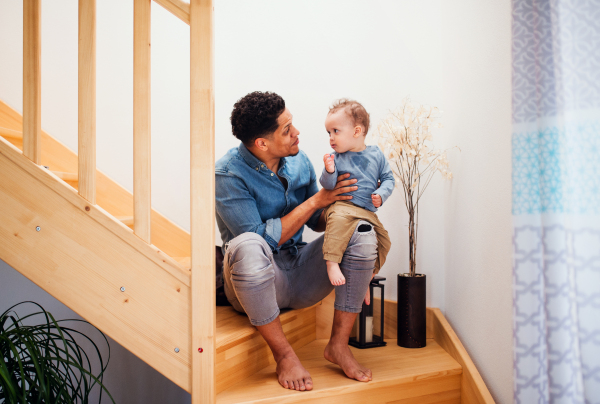 A portrait of father and small toddler son indoors at home, sitting at the bottom of wooden staircase.