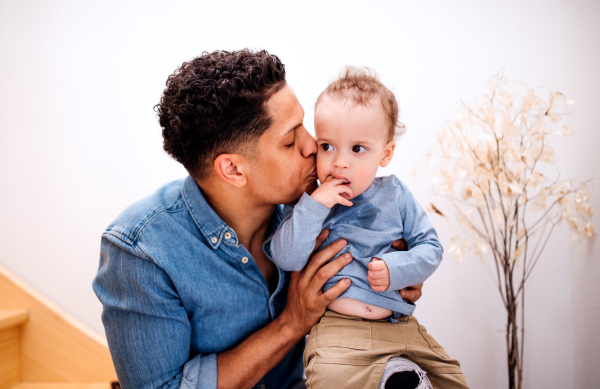 A portrait of father and small toddler son indoors at home, sitting at the bottom of wooden staircase.