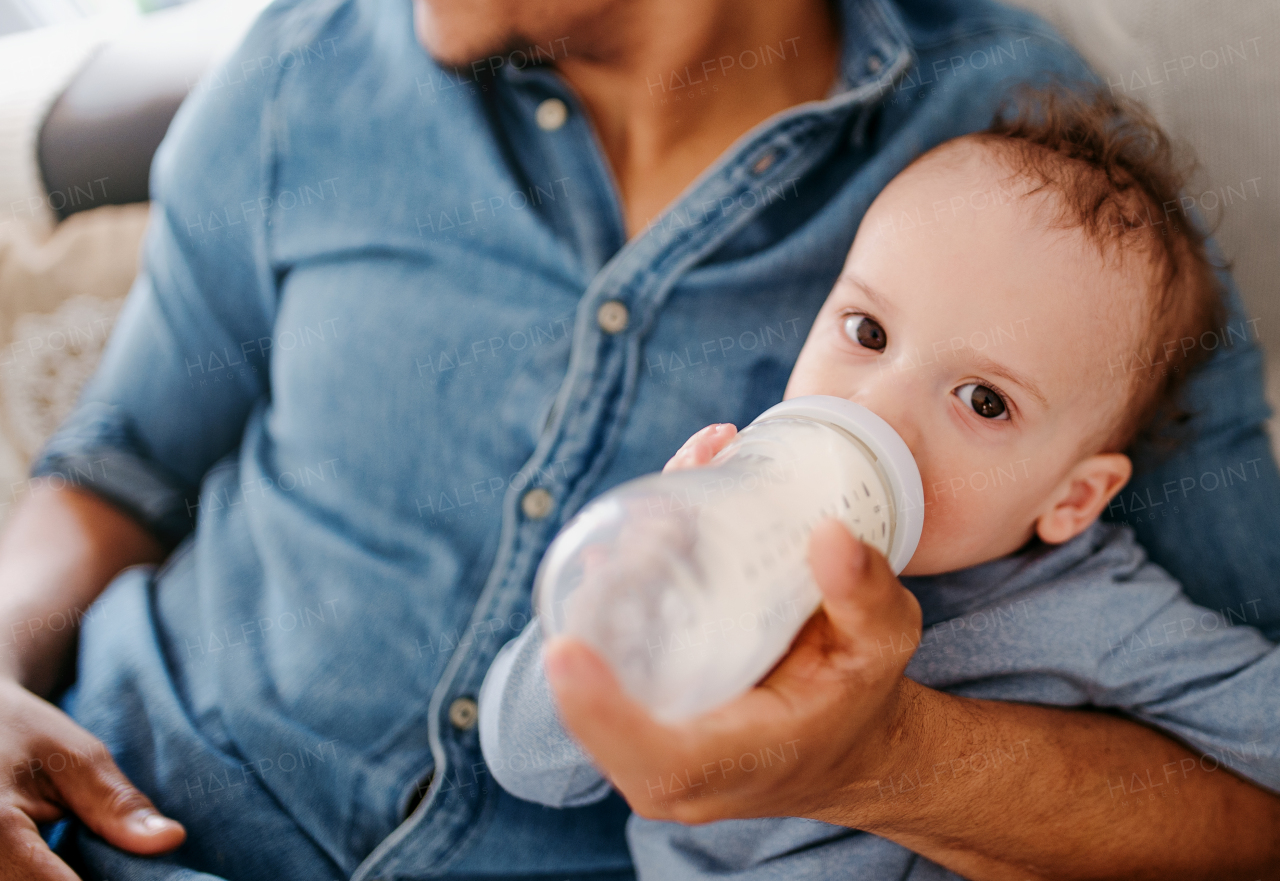 A midsection of unrecognizable father bottle feeding a small toddler son indoors at home.