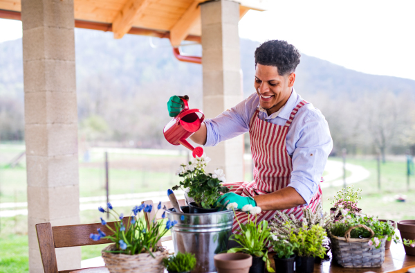 A portrait of young cheerful man gardener outdoors at home, planting and watering flowers.