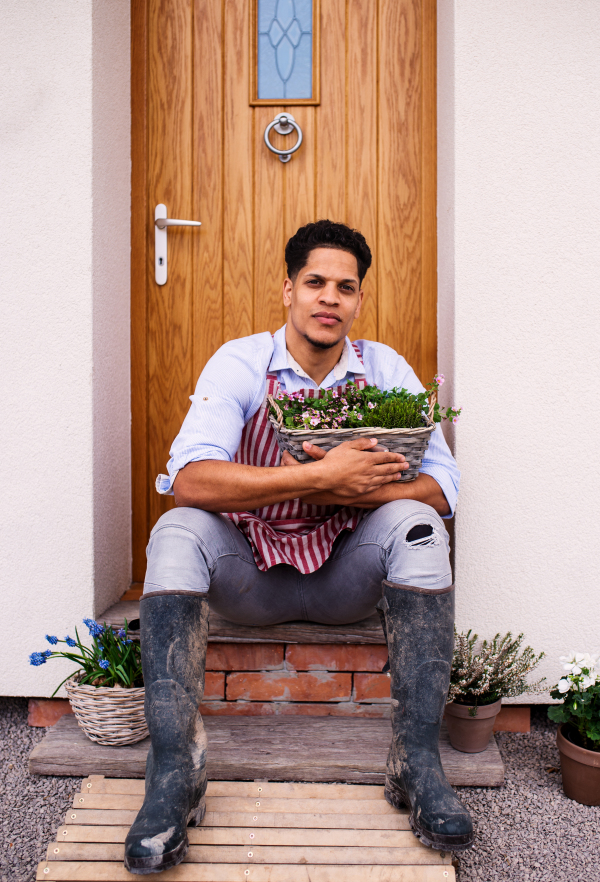 A portrait of young cheerful man gardener sitting in front of door at home, holding basket with flowers.