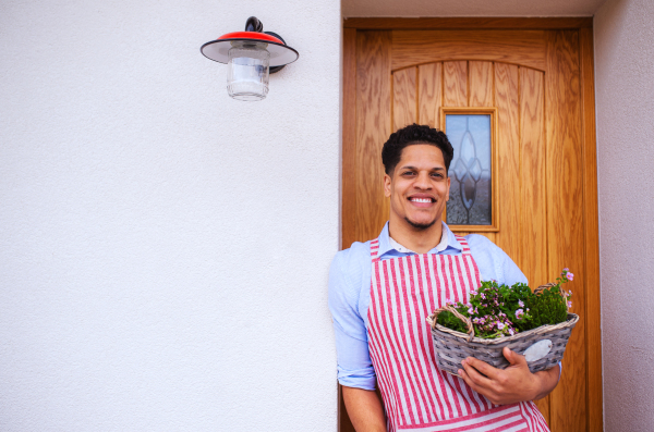 A portrait of young cheerful man gardener standing in front of door at home, holding basket with flowers. Copy space.