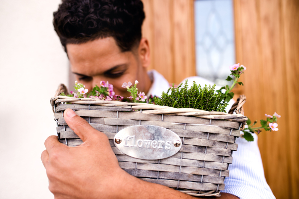 A close-up portrait of young cheerful man gardener standing in front of door at home, holding basket with flowers.