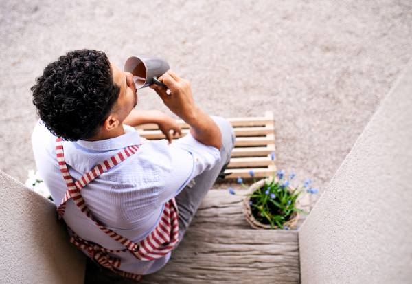 A top view of young man eith apron sitting in front of door at home, drinking tea.