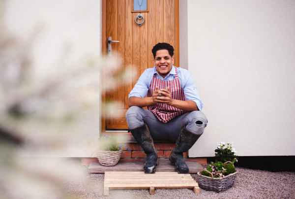 A portrait of young cheerful man gardener sitting in front of door at home, holding coffee.