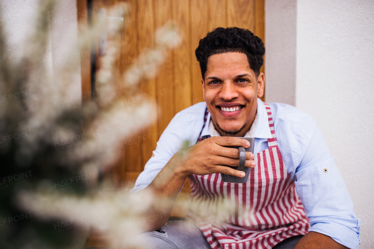 A portrait of young man gardener with coffee sitting in front of door at home.