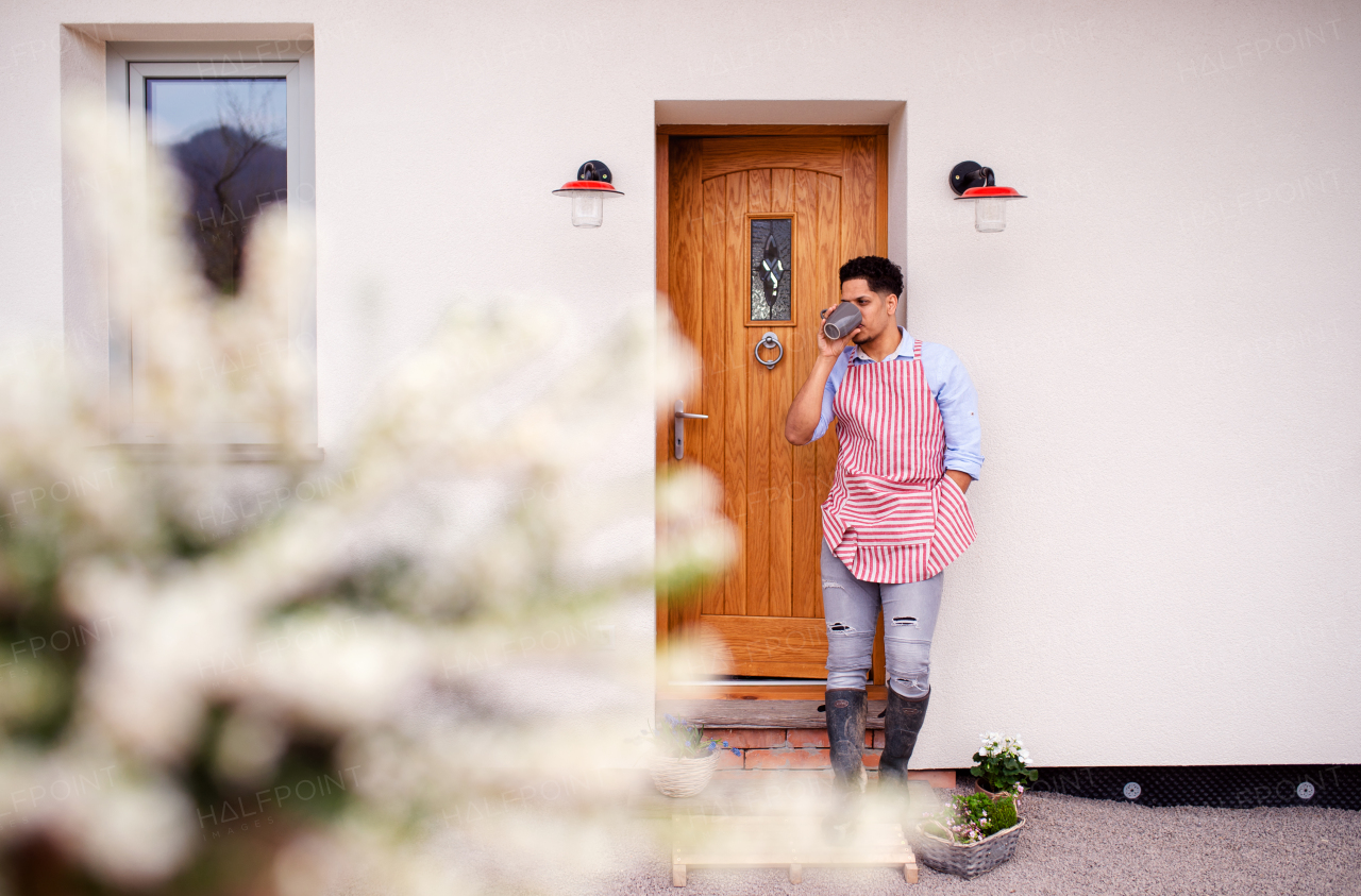 A portrait of young cheerful man gardener standing in front of door at home, holding coffee.