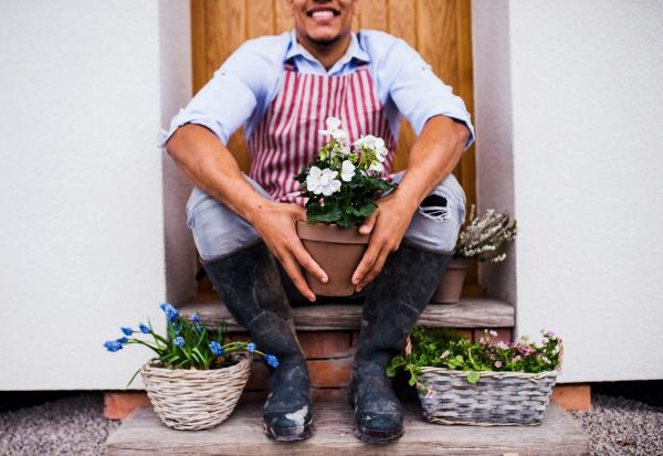 A midsection of unrecognizable young man gardener sitting in front of door at home, holding pot with flowers.