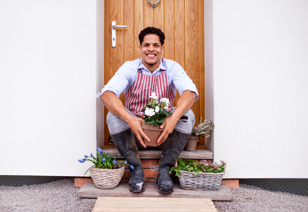 A portrait of young cheerful man sitting in front of door at home, planting flowers.