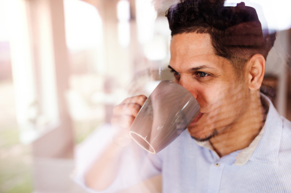 A man indoors standing by window at home, drinking coffee. Shot through glass. Copy space.