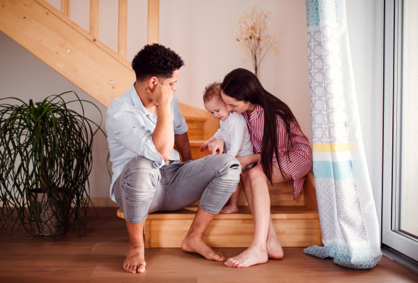 A portrait of young family with small toddler son indoors at home, sitting at the bottom of the stairs.