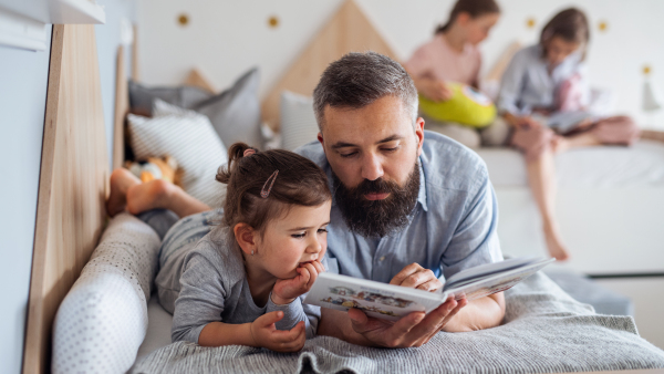 A father with three daughters indoors at home, reading a book.