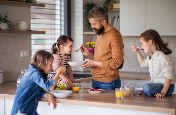 A father with three daughters indoors at home, eating breakfast in kitchen.