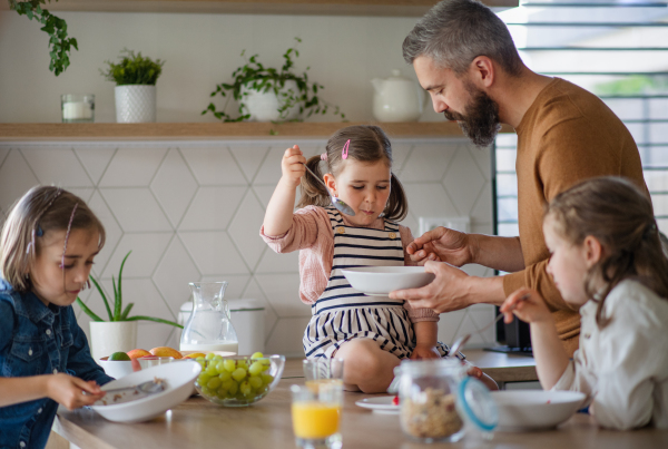 A father with three daughters indoors at home, eating breakfast in kitchen.