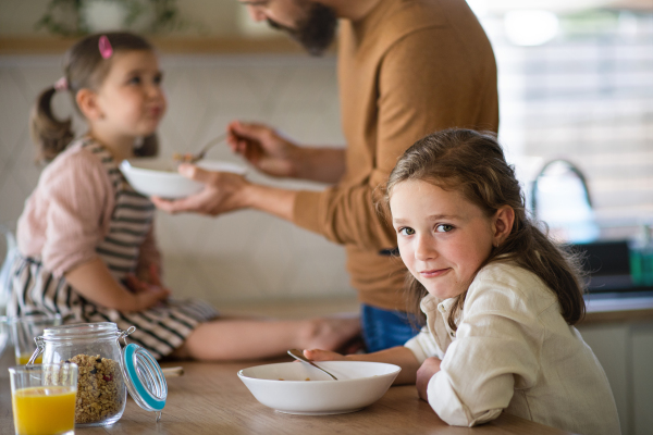 A father with daughters indoors at home, eating breakfast in kitchen.